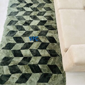 Puede incluir: Una alfombra verde y negra con estampado geom&eacute;trico y textura peluda. La alfombra tiene un borde con flecos y est&aacute; colocada sobre un suelo de baldosas blancas.