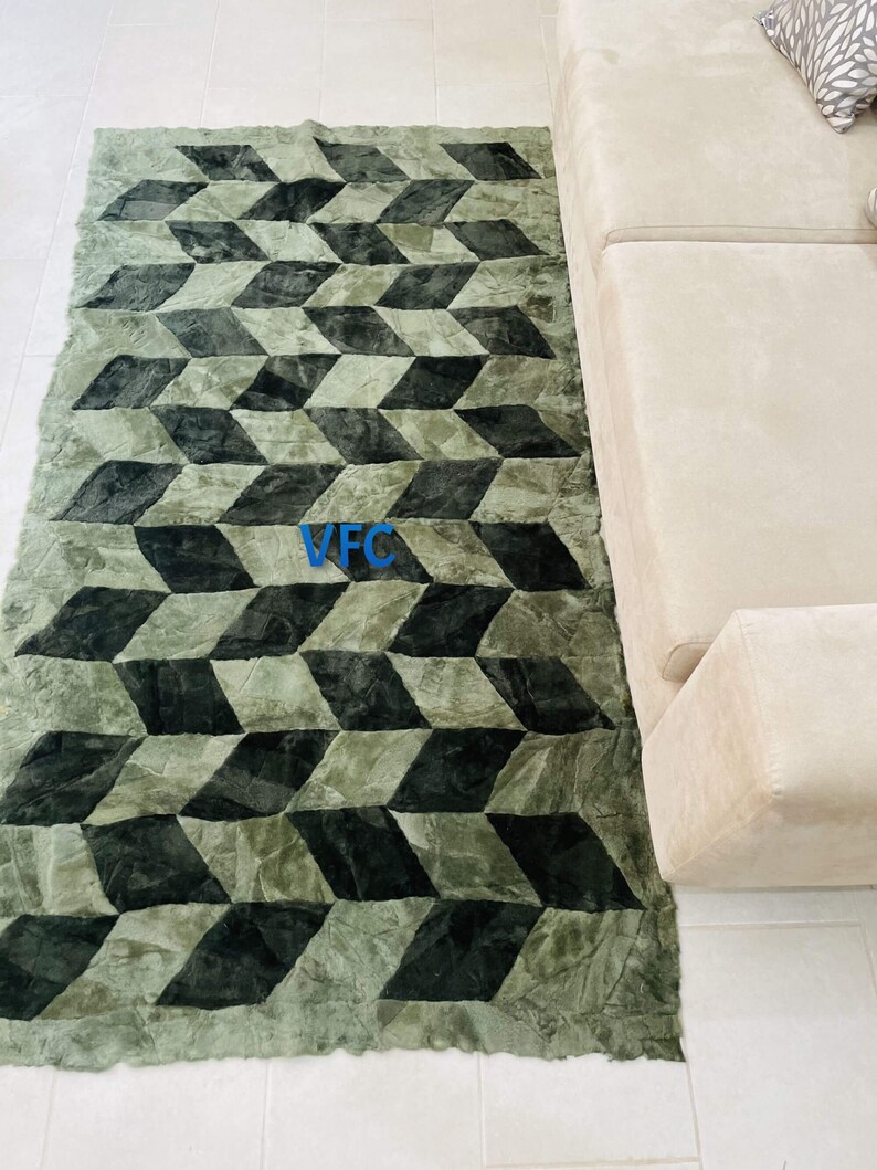 Puede incluir: Una alfombra verde y negra con estampado geom&eacute;trico y textura peluda. La alfombra tiene un borde con flecos y est&aacute; colocada sobre un suelo de baldosas blancas.
