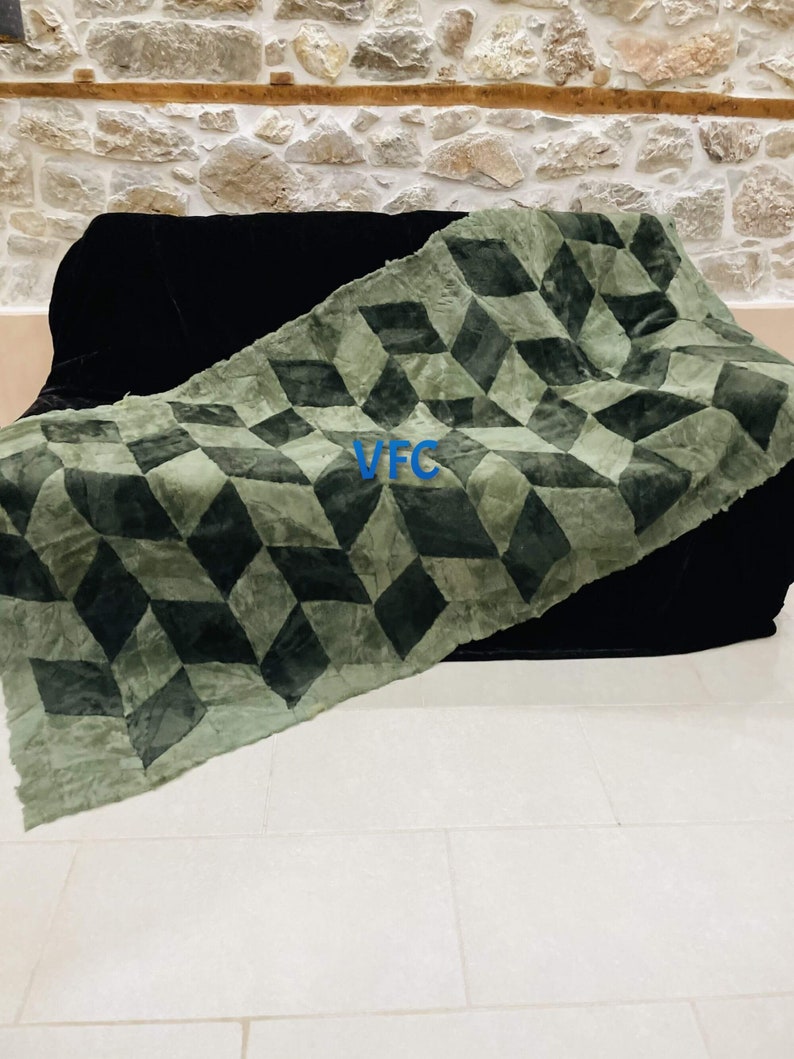 Puede incluir: Una manta de tiro con estampado geom&eacute;trico verde y negro con las letras "VFC" en azul sobre la tela. La manta est&aacute; drapeada sobre un sof&aacute; negro.