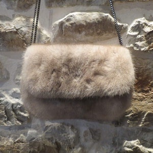 May include: A light brown fur clutch bag with a silver chain strap. The bag is hanging against a stone wall.