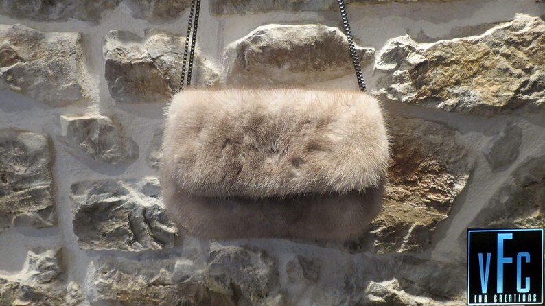 May include: A light brown fur clutch bag with a silver chain strap. The bag is hanging against a stone wall.