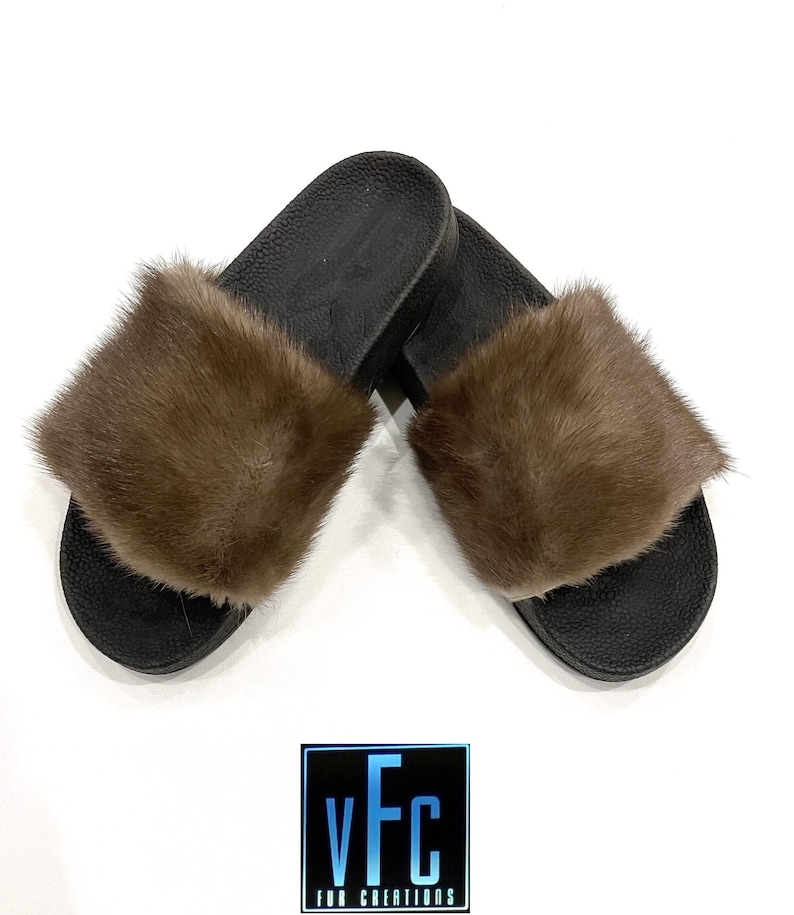 May include: A pair of brown fur slides with black soles. The slides are made of real fur and have a comfortable fit.