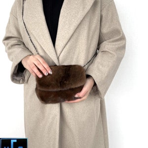 May include: A brown fur crossbody bag with a chain strap. The bag is worn by a person wearing a beige coat.