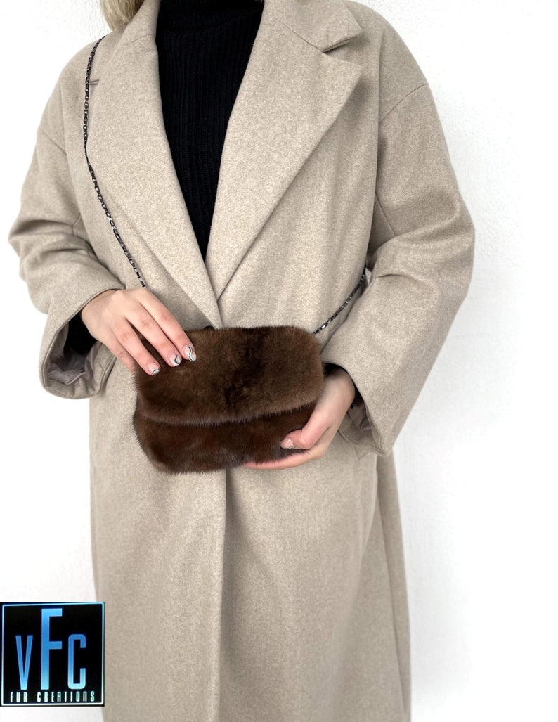 May include: A brown fur crossbody bag with a chain strap. The bag is worn by a person wearing a beige coat.