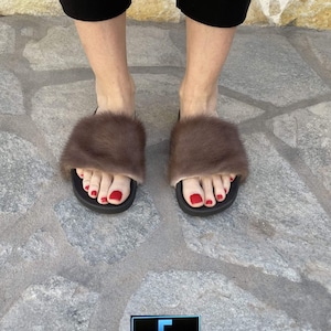 May include: A pair of brown fur slides with a black sole. The slides are worn by a person with red nail polish. The image includes the text "vFc FUR CREATIONS".