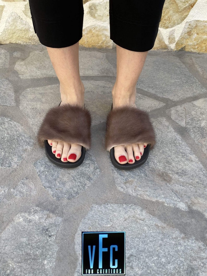 May include: A pair of brown fur slides with a black sole. The slides are worn by a person with red nail polish. The image includes the text "vFc FUR CREATIONS".