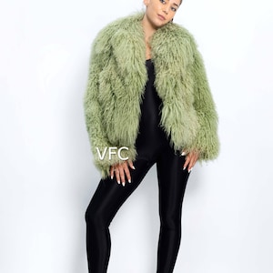 May include: A pale green faux fur jacket with a shaggy texture. The jacket is worn over a black jumpsuit and paired with clear heeled sandals. The image includes the text "VFC".
