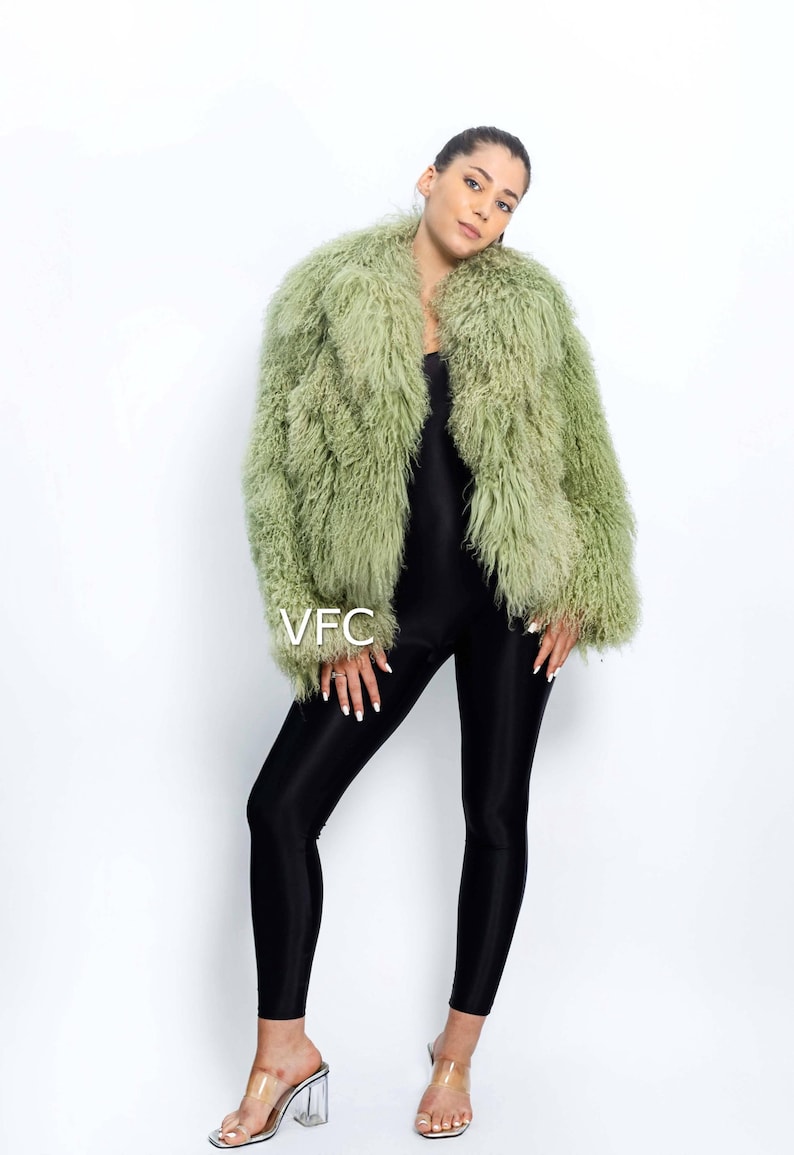 May include: A pale green faux fur jacket with a shaggy texture. The jacket is worn over a black jumpsuit and paired with clear heeled sandals. The image includes the text "VFC".