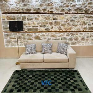 Pode incluir: Um tapete xadrez verde e preto com um logotipo "VFC" no centro. O tapete est&aacute; na frente de um sof&aacute; bege com tr&ecirc;s almofadas. O sof&aacute; est&aacute; na frente de uma parede de pedra com uma l&acirc;mpada.