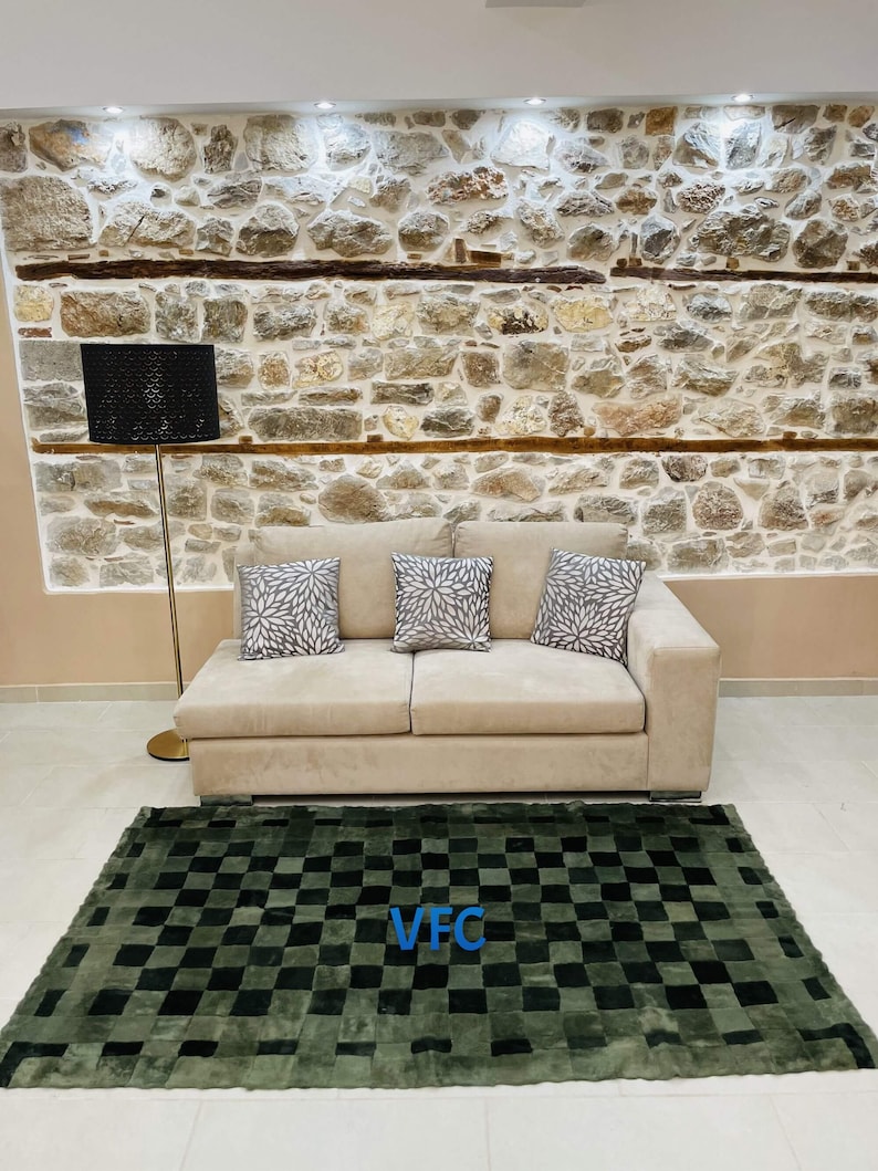 Pode incluir: Um tapete xadrez verde e preto com um logotipo "VFC" no centro. O tapete est&aacute; na frente de um sof&aacute; bege com tr&ecirc;s almofadas. O sof&aacute; est&aacute; na frente de uma parede de pedra com uma l&acirc;mpada.
