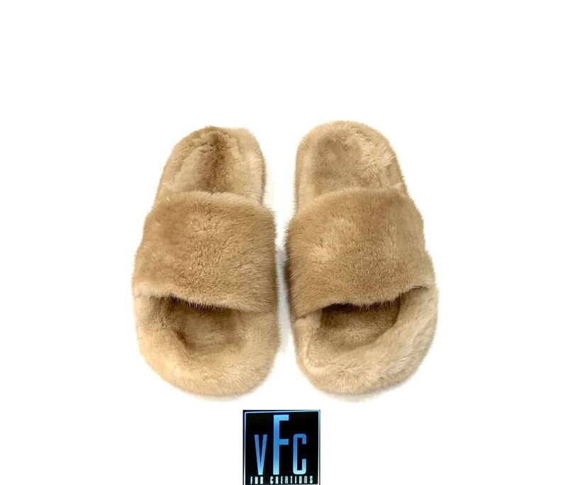 May include: A pair of beige faux fur sliders. The sliders have a wide strap across the top of the foot and a simple, open-toe design.