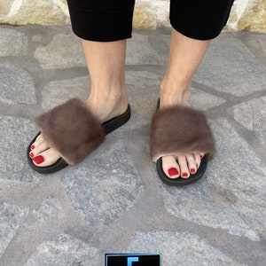 May include: A pair of brown fur slides with black soles. The slides are worn by a person with red nail polish. The background is a stone surface.