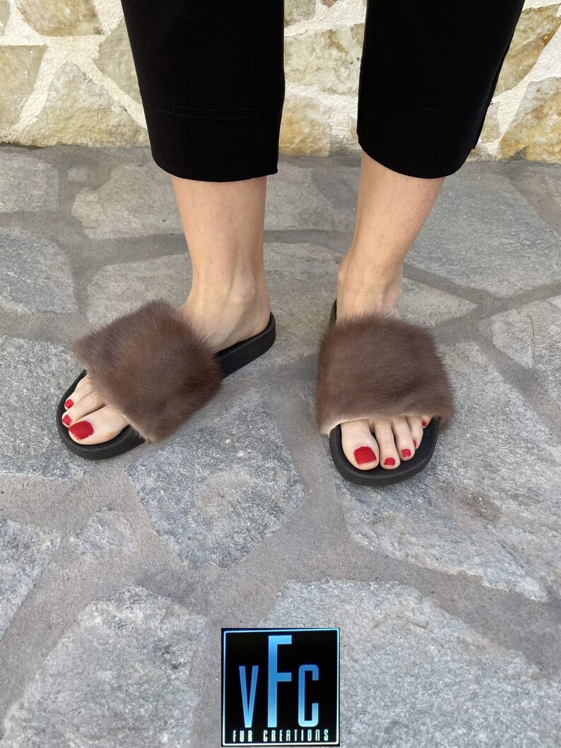 May include: A pair of brown fur slides with black soles. The slides are worn by a person with red nail polish. The background is a stone surface.