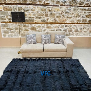 May include: A black faux fur rug with the letters "VFC" in blue text in the center of the rug. The rug is in front of a beige couch with three throw pillows. The couch is in front of a stone wall.