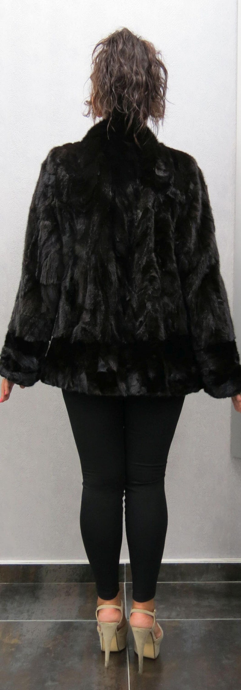 Puede incluir: Un abrigo de piel negro de manga larga y cuello alto. El abrigo se lleva sobre unos leggings negros y tacones altos beige.