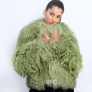 May include: A light green, shaggy jacket with a textured appearance. The jacket has long sleeves and a rounded neckline. The model is wearing black trousers. The jacket is a statement piece.
