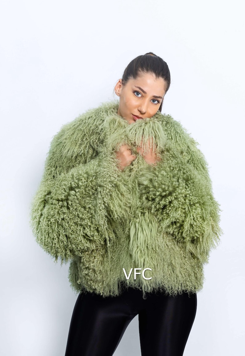 May include: A light green, shaggy jacket with a textured appearance. The jacket has long sleeves and a rounded neckline. The model is wearing black trousers. The jacket is a statement piece.