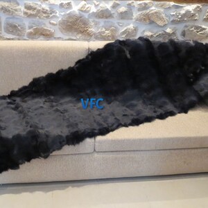 May include: A black sheepskin throw rug is draped over a beige sofa. The rug is long and narrow, with a soft, fluffy texture. The text "VFC" is visible on the rug.