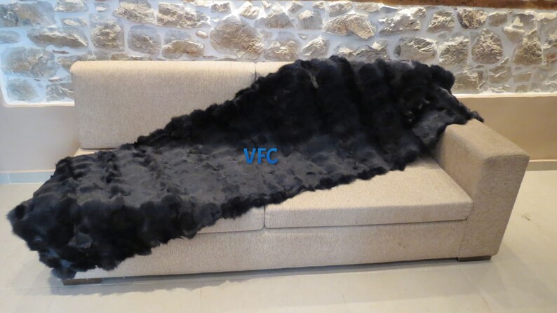 May include: A black sheepskin throw rug is draped over a beige sofa. The rug is long and narrow, with a soft, fluffy texture. The text "VFC" is visible on the rug.