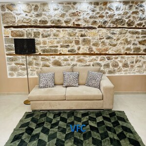 Puede incluir: Un sof&aacute; beige con tres almohadas se encuentra frente a una pared de piedra. Una l&aacute;mpara de pie negra se encuentra junto al sof&aacute;. Una alfombra geom&eacute;trica verde y negra con las letras "VFC" en azul est&aacute; en el suelo frente al sof&aacute;.