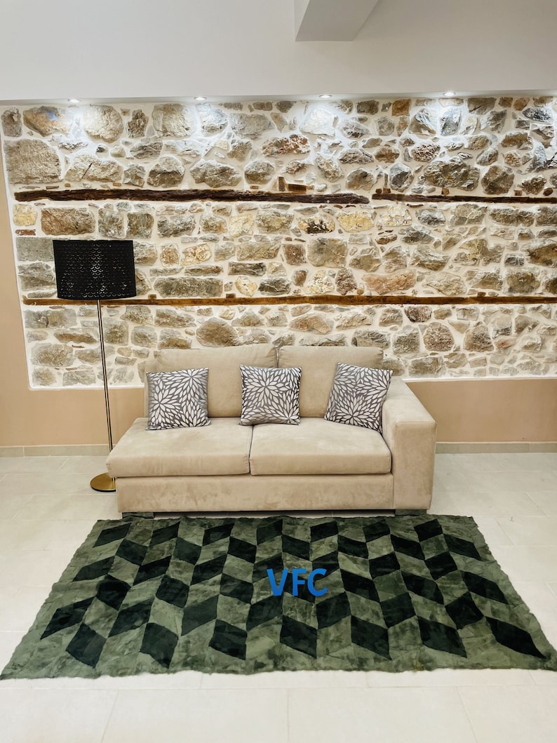 Puede incluir: Un sof&aacute; beige con tres almohadas se encuentra frente a una pared de piedra. Una l&aacute;mpara de pie negra se encuentra junto al sof&aacute;. Una alfombra geom&eacute;trica verde y negra con las letras "VFC" en azul est&aacute; en el suelo frente al sof&aacute;.