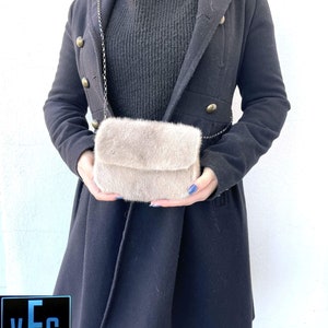 May include: A light brown faux fur crossbody bag with a silver chain strap. The bag is being held by a person wearing a black coat.