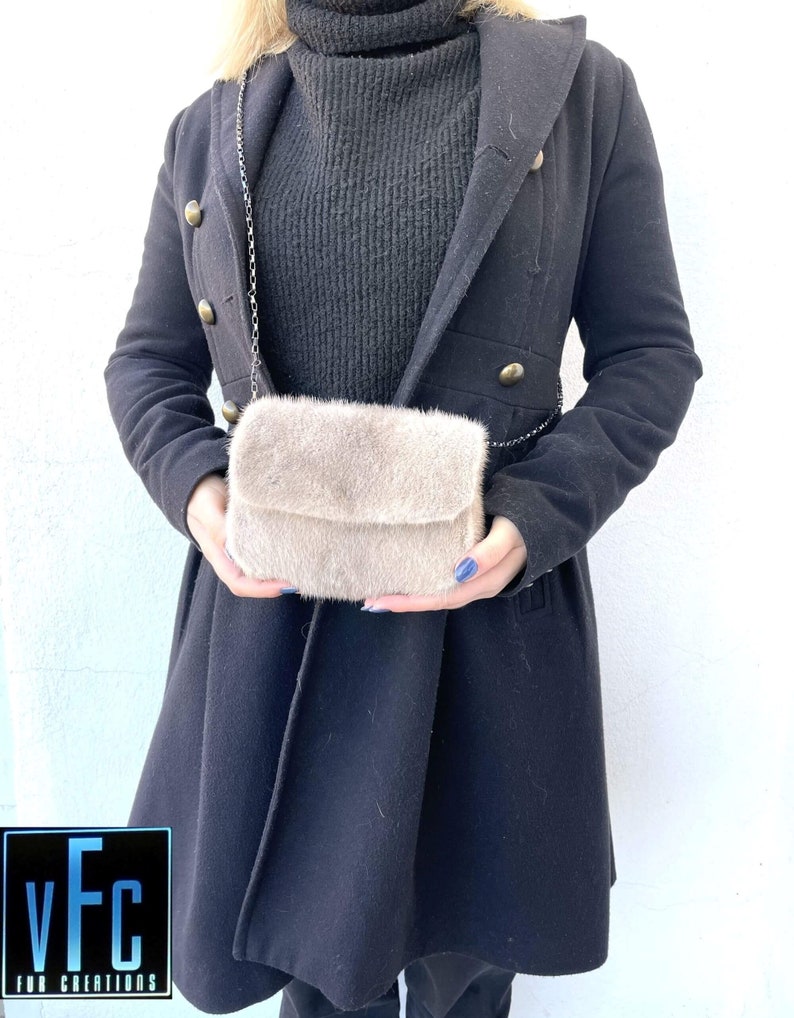 May include: A light brown faux fur crossbody bag with a silver chain strap. The bag is being held by a person wearing a black coat.