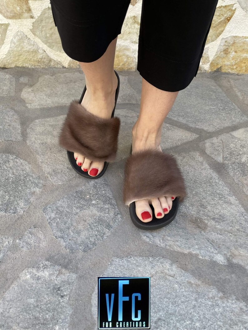 May include: A pair of brown fur slides with black soles. The slides are worn by a person with red nail polish. The background is a stone patio.