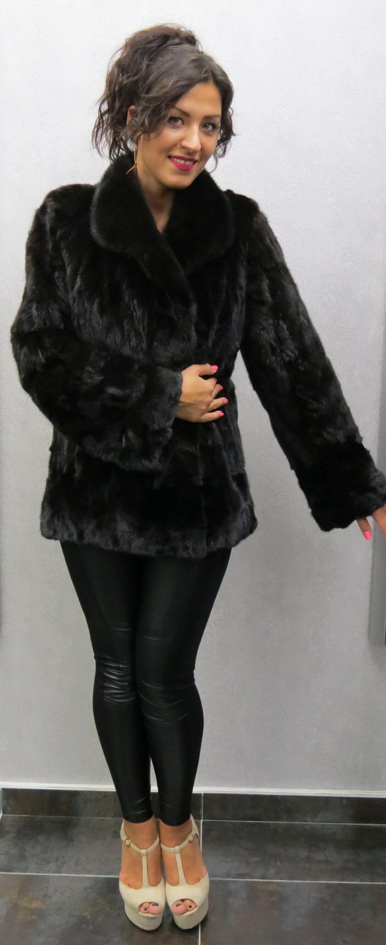 Puede incluir: Un abrigo de piel negro con cuello y mangas largas. La persona que lleva el abrigo tambi&eacute;n lleva leggings negros y tacones beige.
