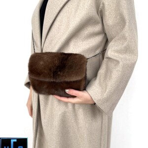 May include: A brown fur crossbody bag with a chain strap. The bag is being held by a person wearing a beige coat.