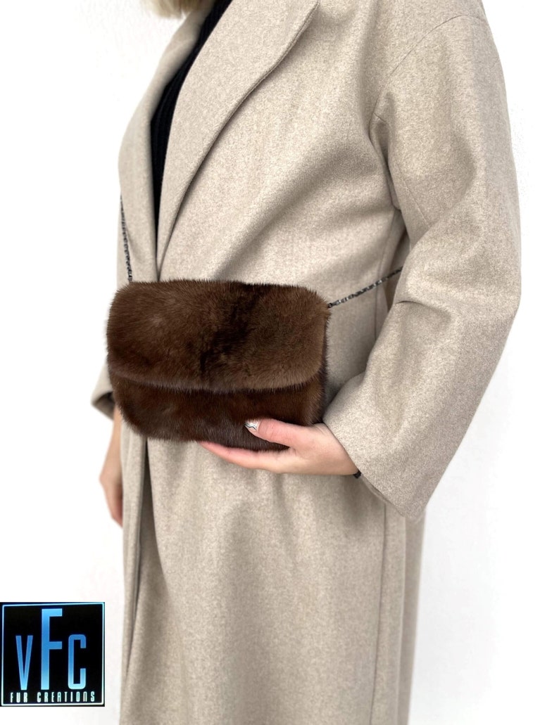 May include: A brown fur crossbody bag with a chain strap. The bag is being held by a person wearing a beige coat.