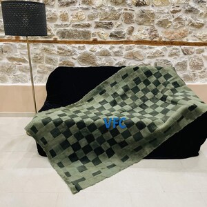 Pode incluir: Uma manta de l&atilde; xadrez verde e preta com o texto "VFC" em letras azuis. A manta est&aacute; colocada sobre um sof&aacute; preto em frente a uma parede de pedra.