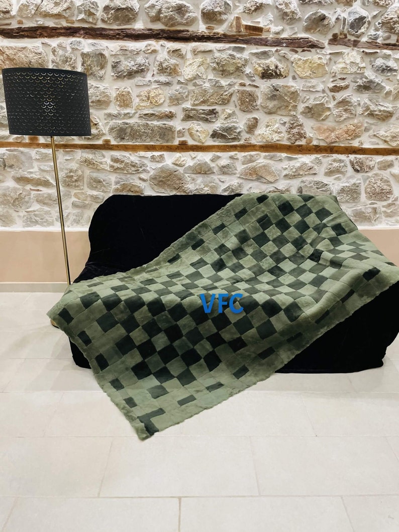Pode incluir: Uma manta de l&atilde; xadrez verde e preta com o texto "VFC" em letras azuis. A manta est&aacute; colocada sobre um sof&aacute; preto em frente a uma parede de pedra.