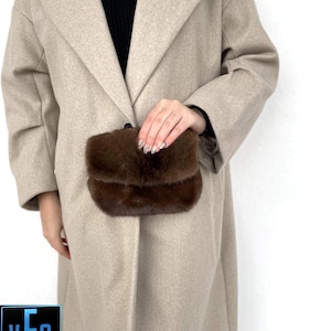 May include: A brown fur clutch purse with a flap closure. The purse is being held by a person wearing a beige coat.