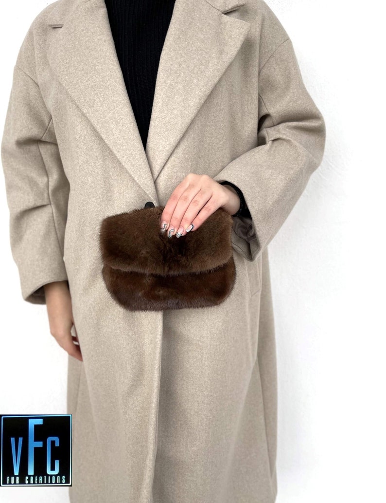 May include: A brown fur clutch purse with a flap closure. The purse is being held by a person wearing a beige coat.