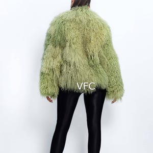 May include: A light green faux fur jacket with a textured appearance. The jacket has a round collar and long sleeves. The model is wearing black leggings and clear heeled sandals. The text "VFC" is visible on the jacket.