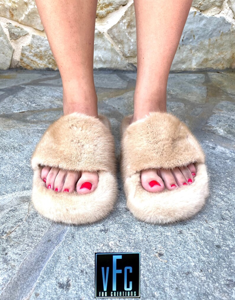 May include: A pair of beige fur slides with a fluffy texture. The slides have a simple design and are perfect for casual wear.