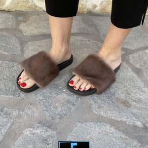 May include: A pair of brown fur slides with black soles. The slides are worn by a person with red nail polish. The image includes the text "vFC FUR CREATIONS".