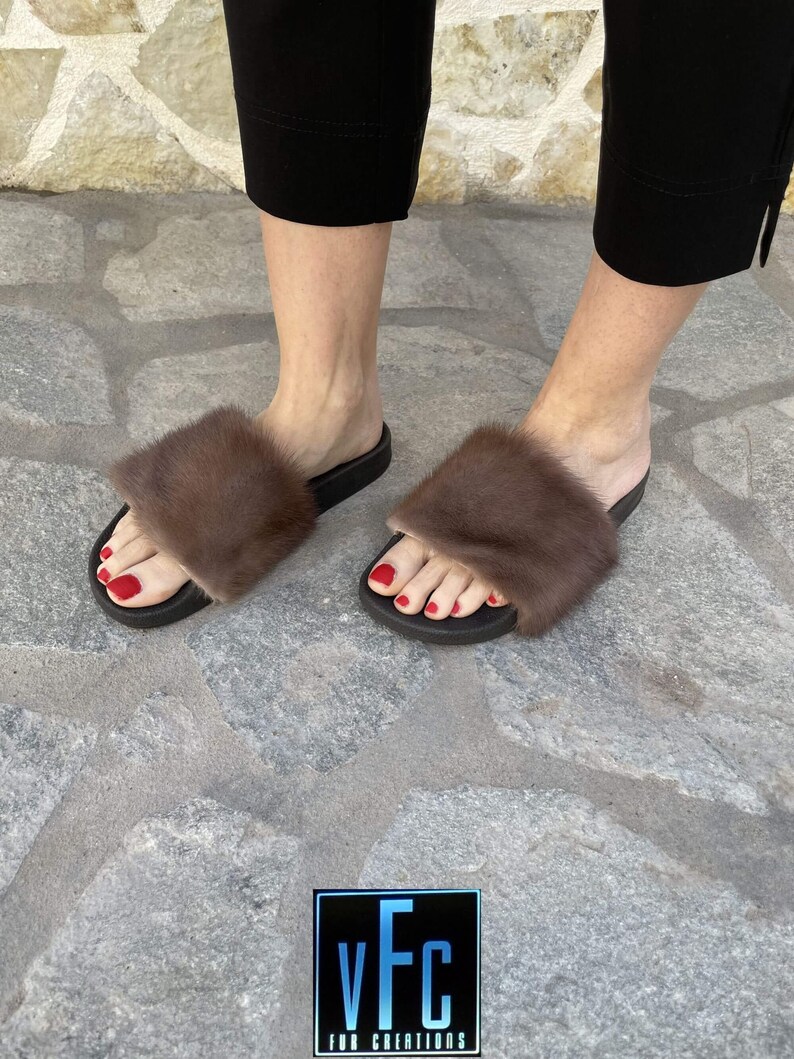 May include: A pair of brown fur slides with black soles. The slides are worn by a person with red nail polish. The image includes the text "vFC FUR CREATIONS".