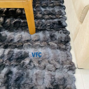 May include: A black and gray fur rug with the letters "VFC" printed in white on the rug. The rug is laid out on a light colored floor and a wooden table is partially visible in the top left corner of the image.