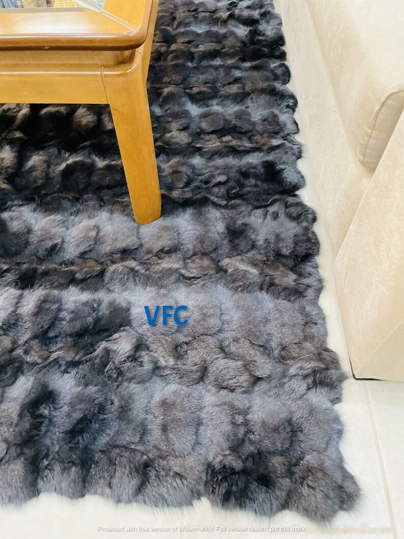 May include: A black and gray fur rug with the letters "VFC" printed in white on the rug. The rug is laid out on a light colored floor and a wooden table is partially visible in the top left corner of the image.