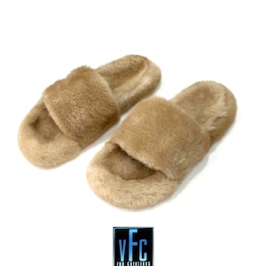 May include: A pair of beige fur slides. The slides are made of soft, fluffy fur and have a simple, slip-on design.