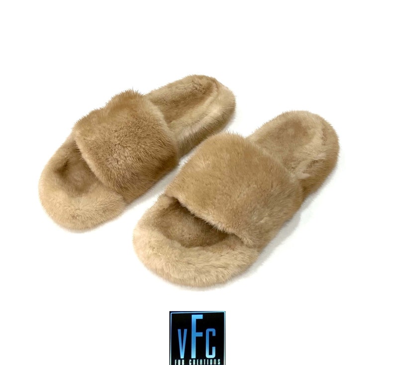 May include: A pair of beige fur slides. The slides are made of soft, fluffy fur and have a simple, slip-on design.