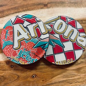 May include: Two round Arizona Iced Tea coasters with a red, white, and blue checkered pattern. One coaster features the Arizona logo with the text "Arizona Iced Tea with Raspberry" and the other coaster features the text "Natural".