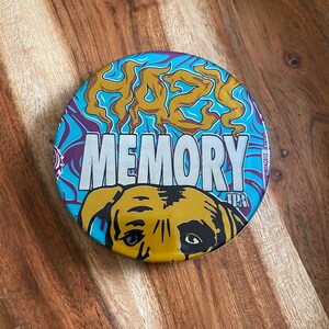 May include: A round, blue, purple, and gold coaster with a dog's face and the words "Haze Memory IPA" printed on it.