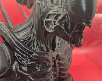 May include: A black, 3D-printed bust of an Alien creature. The sculpture features a detailed head with a smooth, elongated skull, and intricate facial features. The bust is mounted on a circular base, showcasing the creature's upper body.