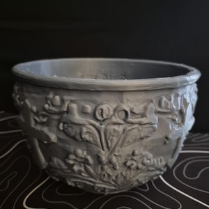 May include: A gray, decorative planter with a detailed floral relief pattern. The planter has a wide, rounded shape and a thick rim. The relief design features flowers and leaves, adding a touch of elegance to the piece. The background is black.