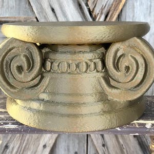 May include: A gold-colored, decorative Ionic column capital. The capital features a scrolled volute design on either side, with a detailed band of egg-and-dart molding. The base is a simple, rounded shape.