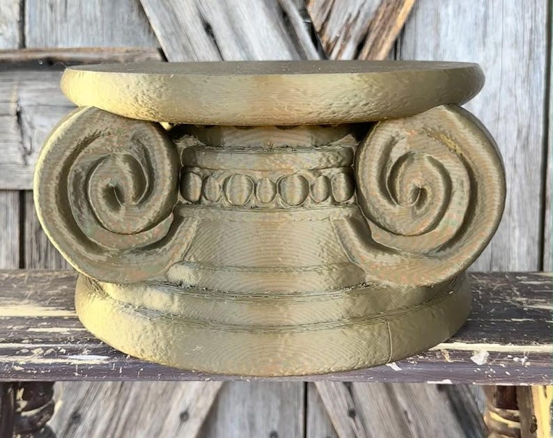 May include: A gold-colored, decorative Ionic column capital. The capital features a scrolled volute design on either side, with a detailed band of egg-and-dart molding. The base is a simple, rounded shape.