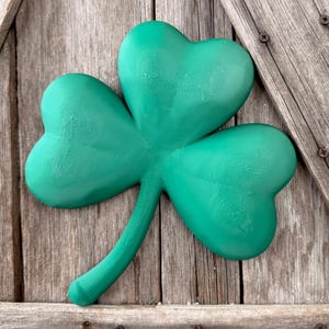 May include: A vibrant green shamrock, a symbol of St. Patrick's Day, is displayed against a weathered wooden background. The three-leaf clover is painted in a solid, bright green color, with a detailed texture. The stem is curved and extends downward.